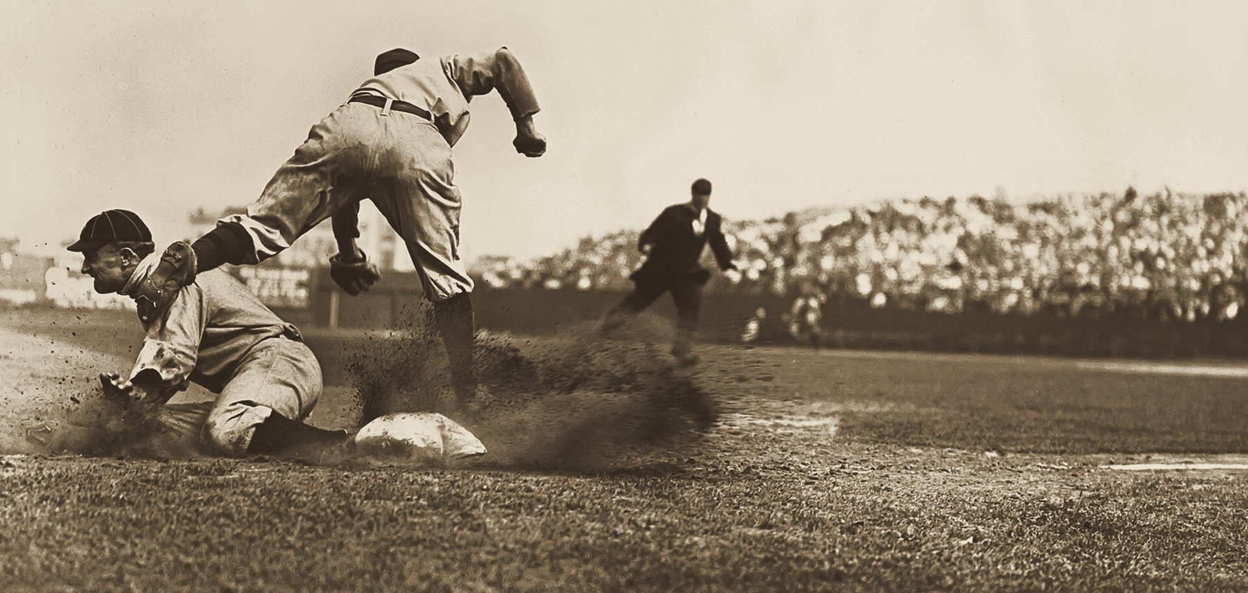 Ty Cobb slides into third base in a cloud of dust as the third baseman leaps over him, with a packed grandstand behind, 1909.